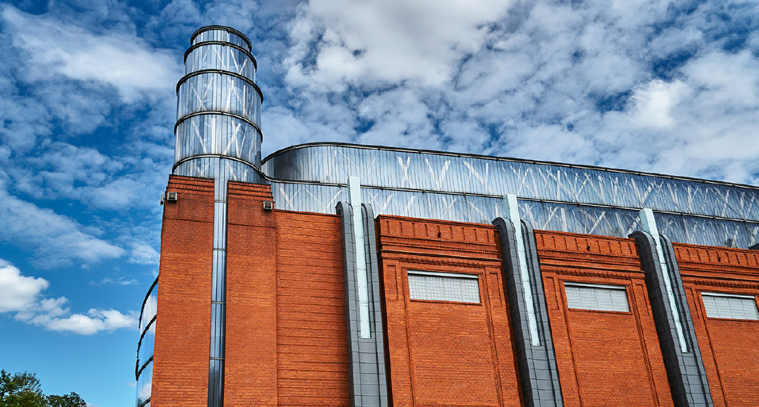 Upwards view of a redbrick large building, with a metal tower and panels.